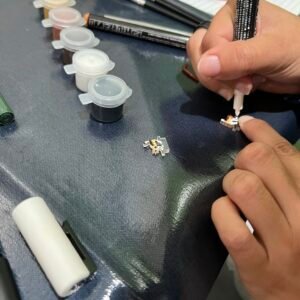 Close-up of an artist painting two laser-cut clear acrylic cat pieces on a worktable, using a fine paintbrush and a paint marker. Small paint pots and tools are arranged nearby as the acrylic pieces are transformed into a calico cat and an orange tabby cat during a hands-on art session.