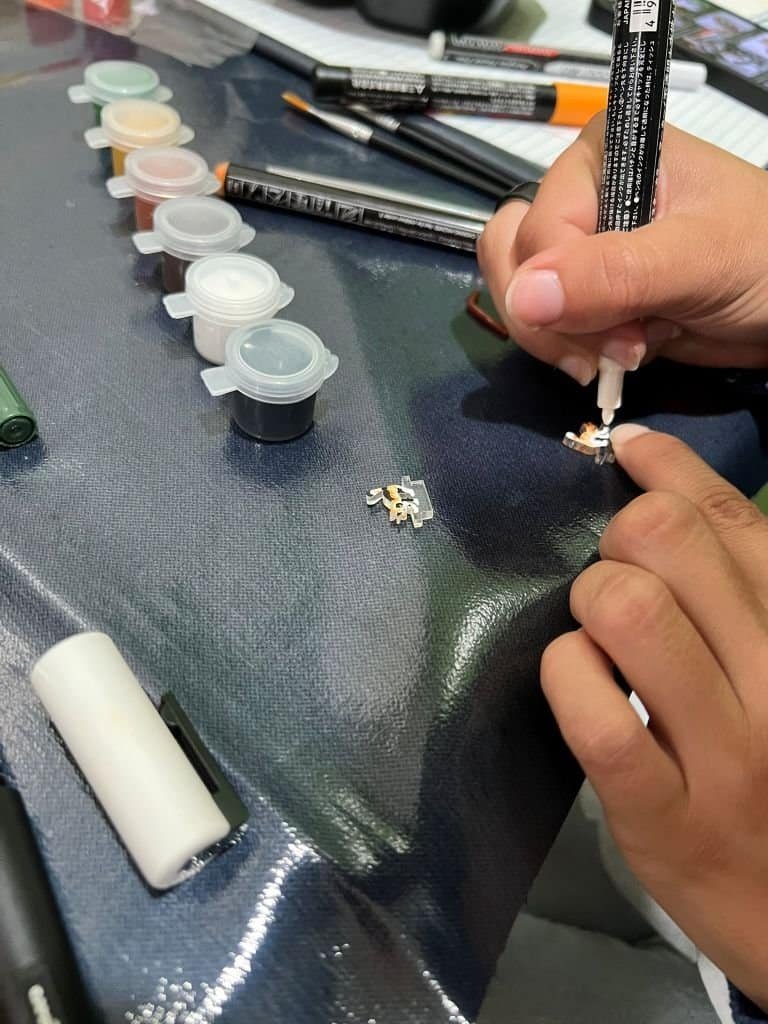 Close-up of an artist painting two laser-cut clear acrylic cat pieces on a worktable, using a fine paintbrush and a paint marker. Small paint pots and tools are arranged nearby as the acrylic pieces are transformed into a calico cat and an orange tabby cat during a hands-on art session.