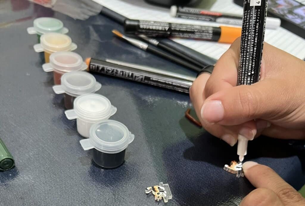 Close-up of an artist painting two laser-cut clear acrylic cat pieces on a worktable, using a fine paintbrush and a paint marker. Small paint pots and tools are arranged nearby as the acrylic pieces are transformed into a calico cat and an orange tabby cat during a hands-on art session.