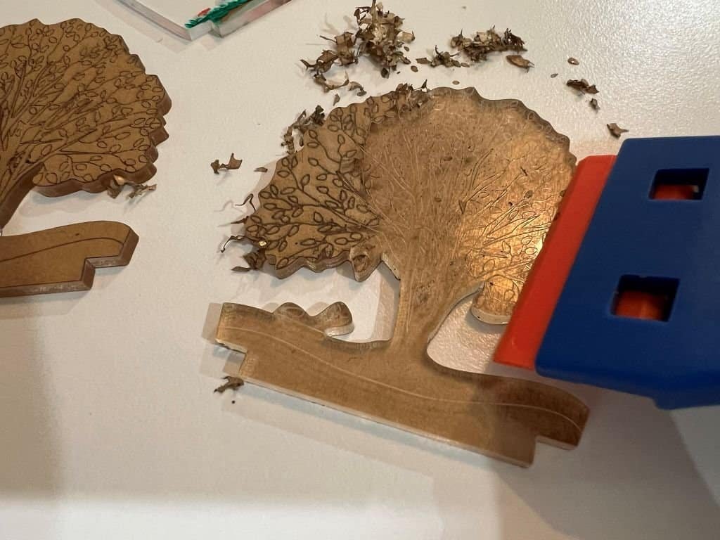 A partially cleaned engraved acrylic tree design shown with paper crumbs on and around it and orange razor sitting on top .